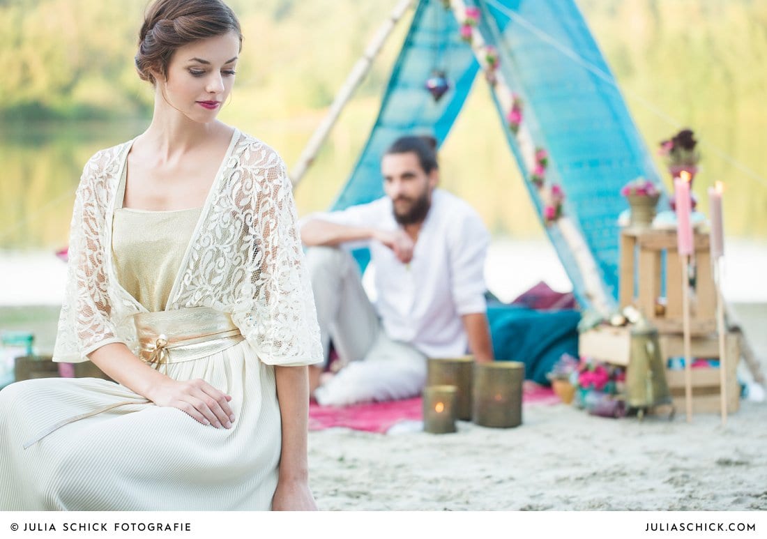 Braut im goldenen Spitzenbrautkleid sitzend vor Boho-Zelt mit Bräutigam im Zelt