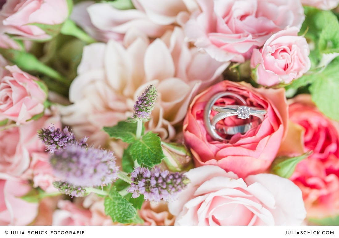 Detailaufnahme von Eheringen und Verlobungsring auf rosa-pfirsichfarbenen Brautstrauß mit Rosen, Nelken und Minze