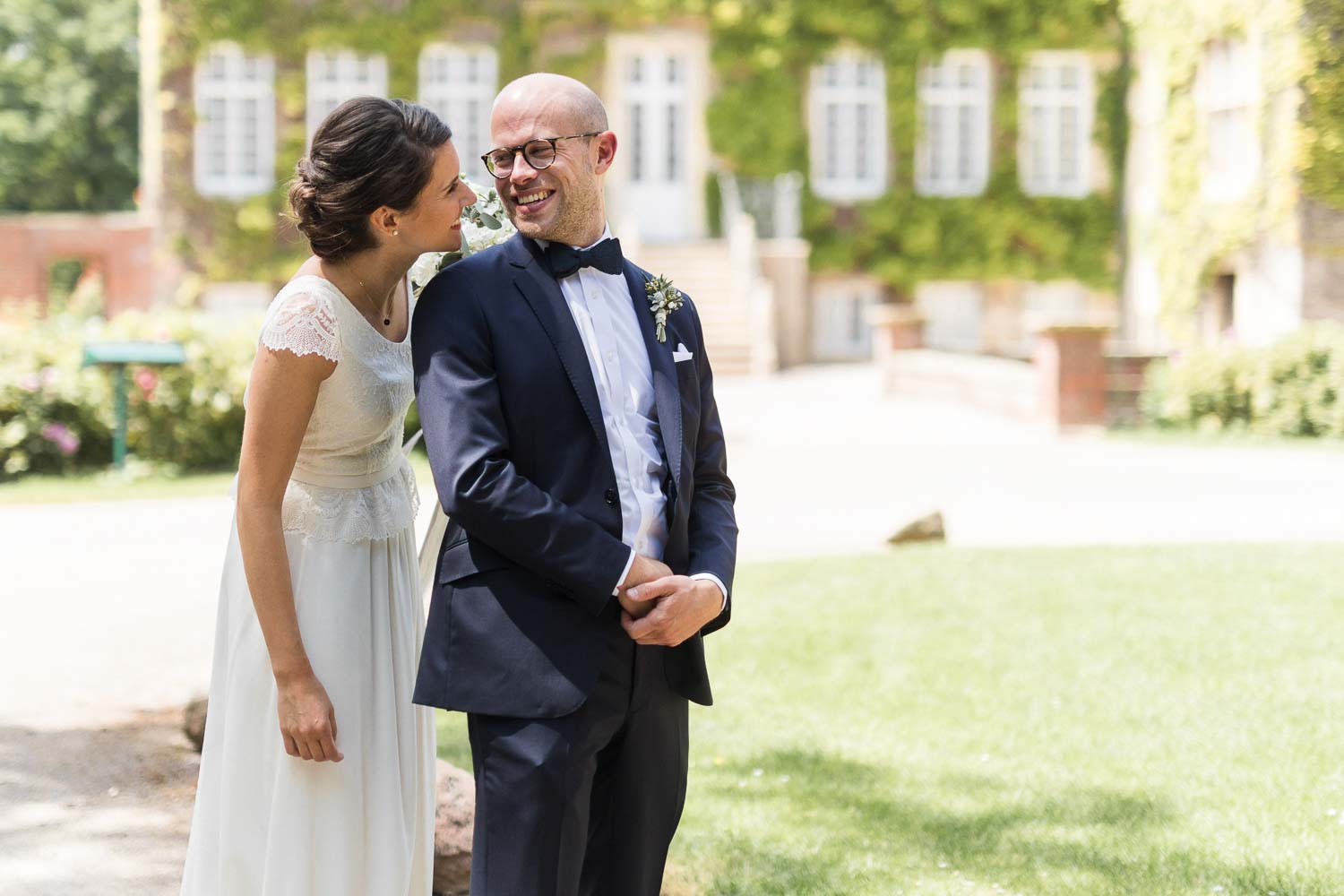 Bräutigam schaut beim First Look über seine Schulter ins Gesicht seiner strahlenden Braut. Im Hintergrund ist die Hochzeitslocation Haus Ruhr zu sehen.