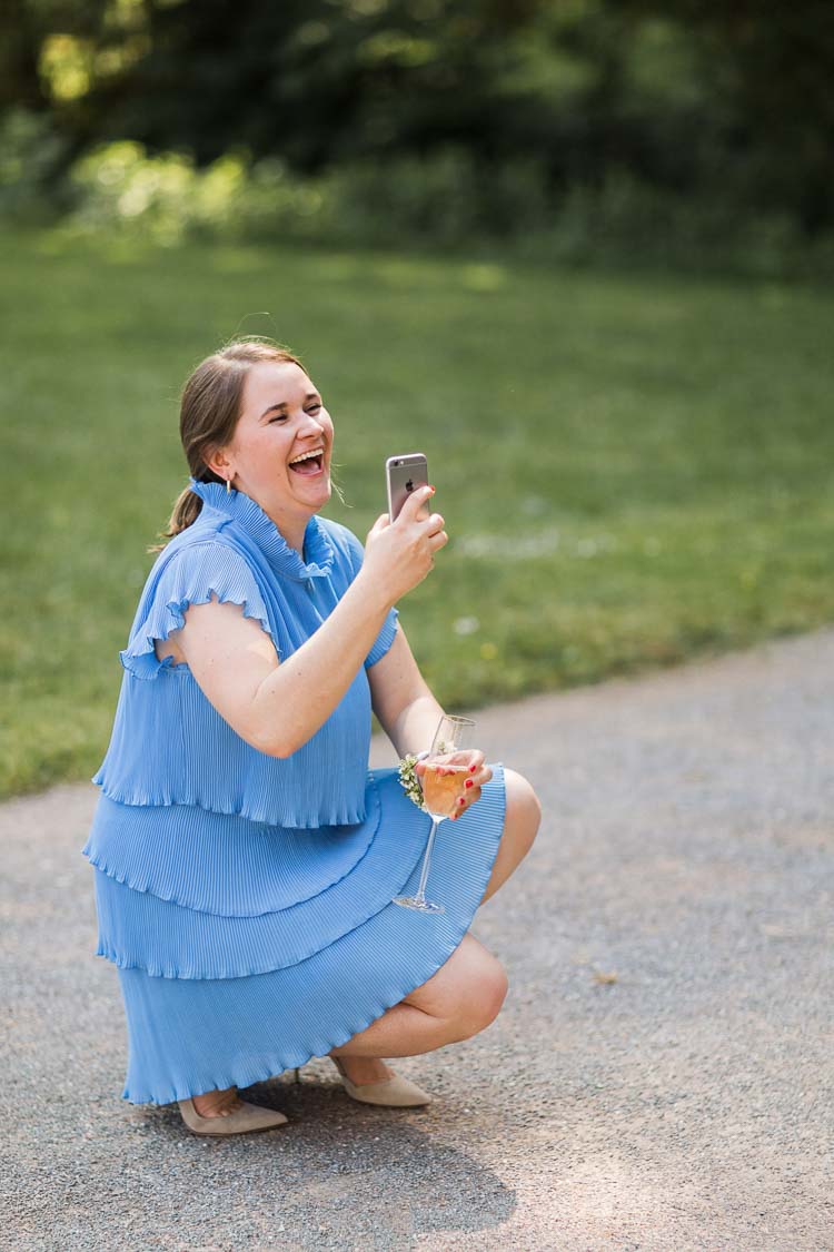 Hochzeitsgast in blauem Kleid macht lachend ein Foto im Garten von Haus Ruhr