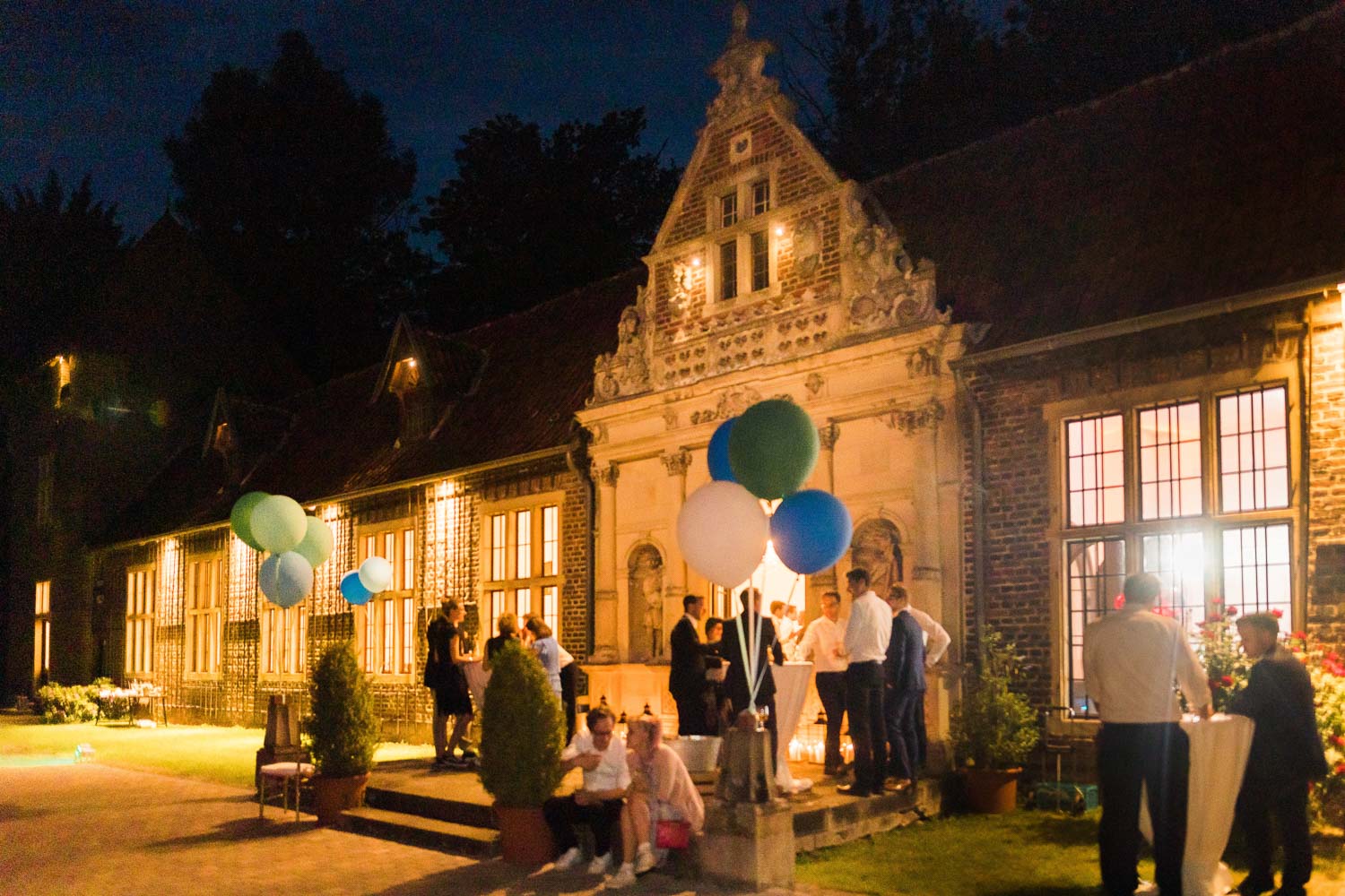 Gäste stehen an Stehtischen vor der Orangerie von Haus Ruhr in Senden bei einer Hochzeitsfeier während der blauen Stunde. Die Orangerie ist hell erleuchtet mit Kerzen und Luftballons.