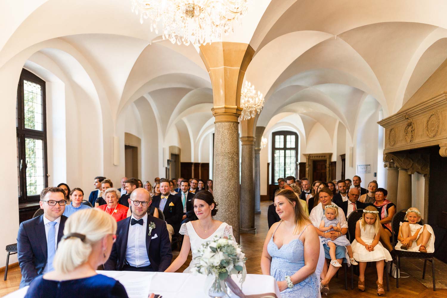 Braut und Bräutigam sowie Trauzeugen bei standesamtlicher Trauung im Rittersaal von Haus Ruhr sitzen am Trautisch. Im Vordergrund sitzt die Standesbeamtin Oda von und zur Mühlen, im Hintergrund sitzen die Gäste