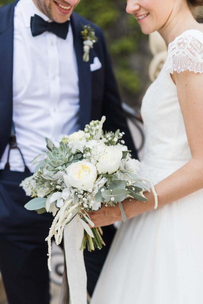 Brautstrauß in weiß und Creme mit Pfingstrosen, Eukalyptus und Echeverien, Braut und Bräutigam im Hintergrund