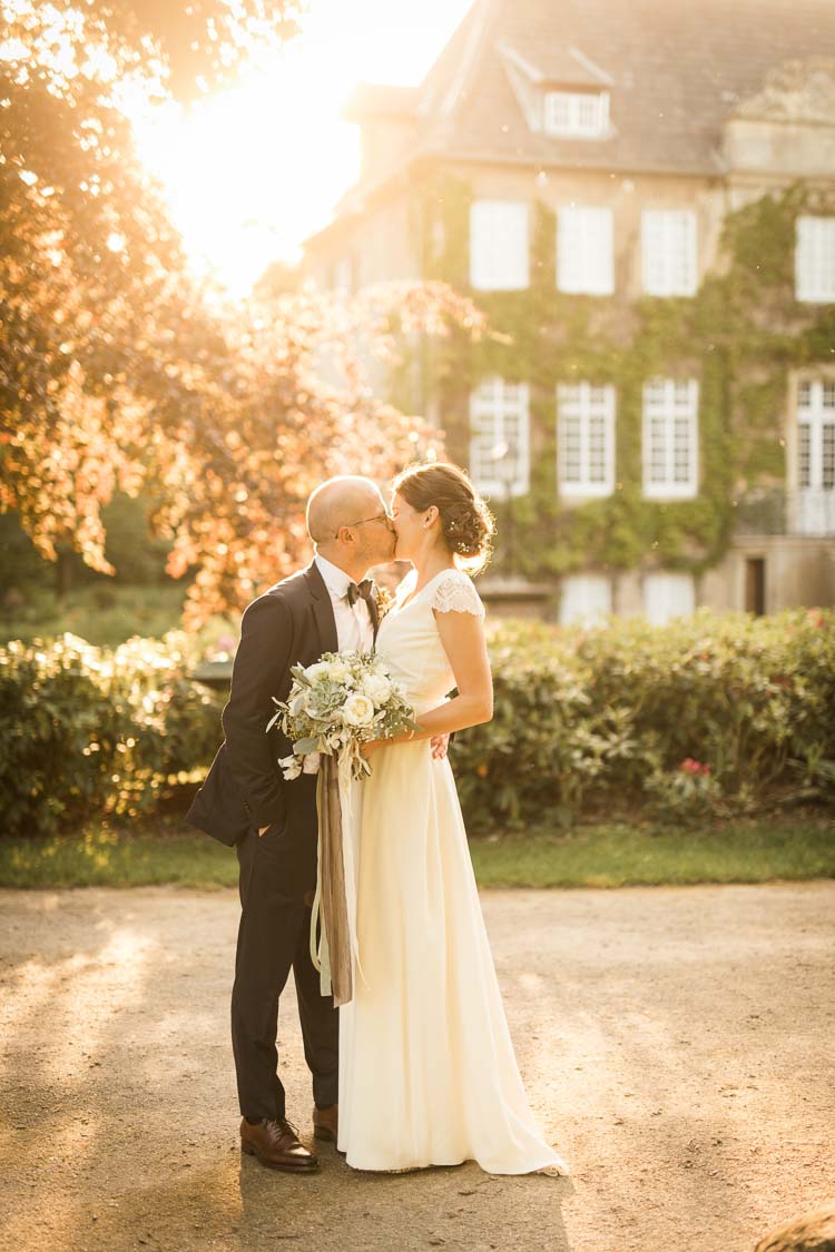 Hochzeitsfotos während der golden Hour. Im Vordergrund küsst sich das Brautpaar glücklich an. Im Hintergrund ist die Hochzeitslocation Haus Ruhr zu sehen.