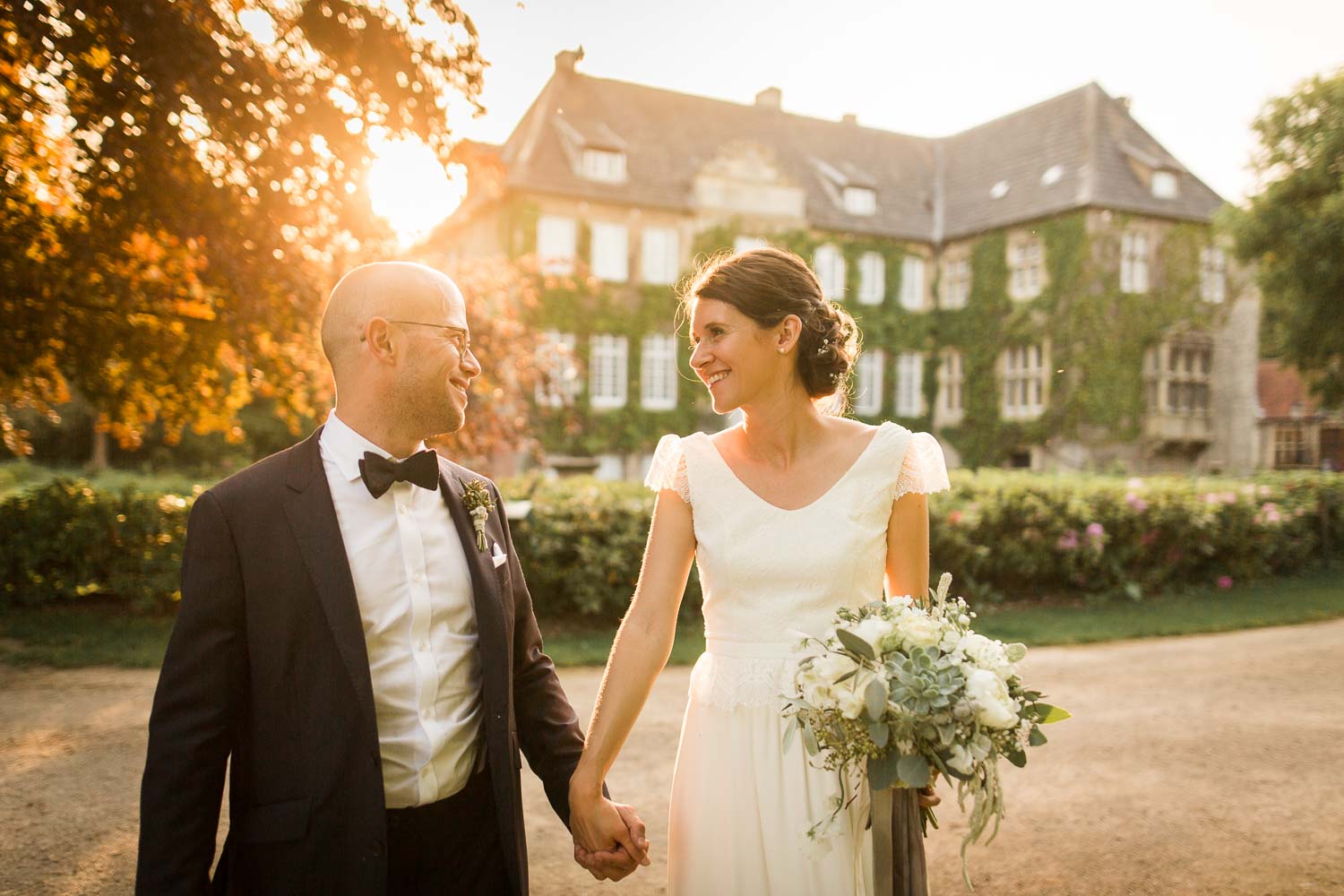 Hochzeitsfotos während der golden Hour. Im Vordergrund lächelt sich das Brautpaar glücklich an. Im Hintergrund ist die Hochzeitslocation Haus Ruhr zu sehen.