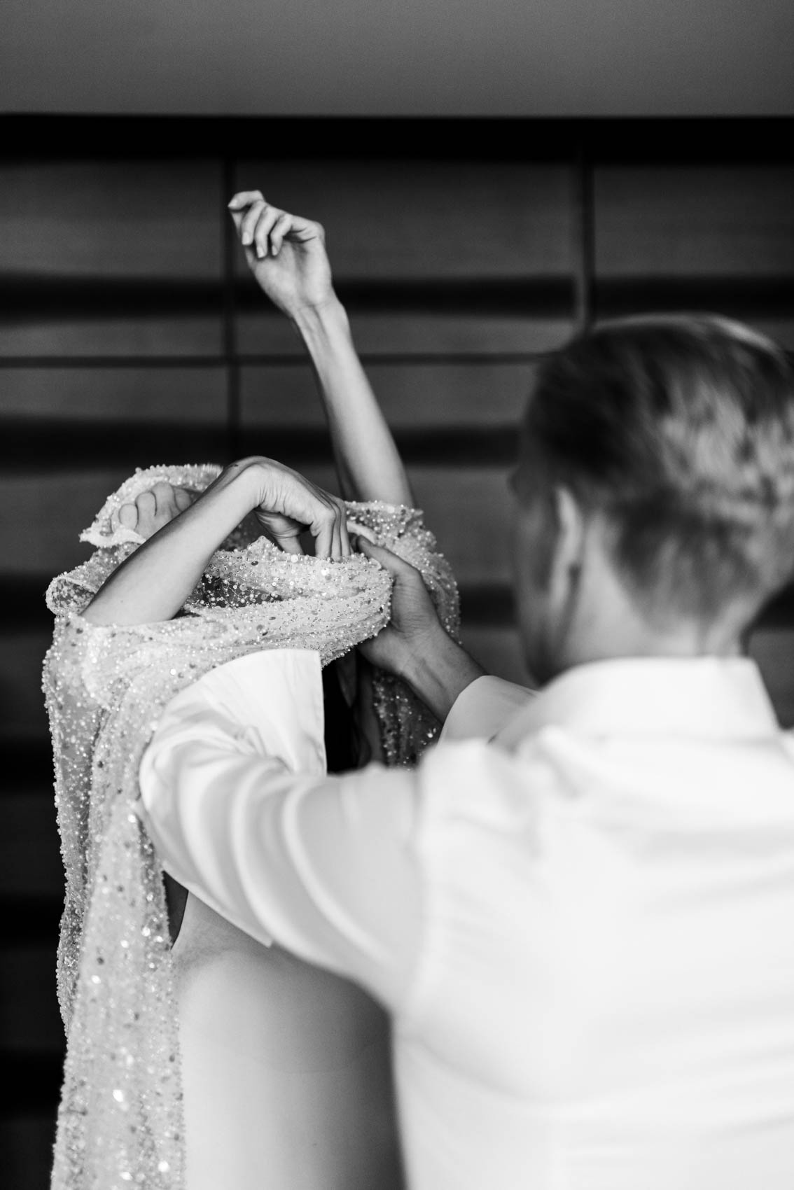 Hochzeitsfoto in schwarz-weiß. Braut hat die Arme in die Luft gestreckt während der Bräutigam ihr bei Anziehen eines Überkleides über das Brautkleid hilft. Das Brautpaar befindet sich in einer Suite des Factory Hotels in Münster