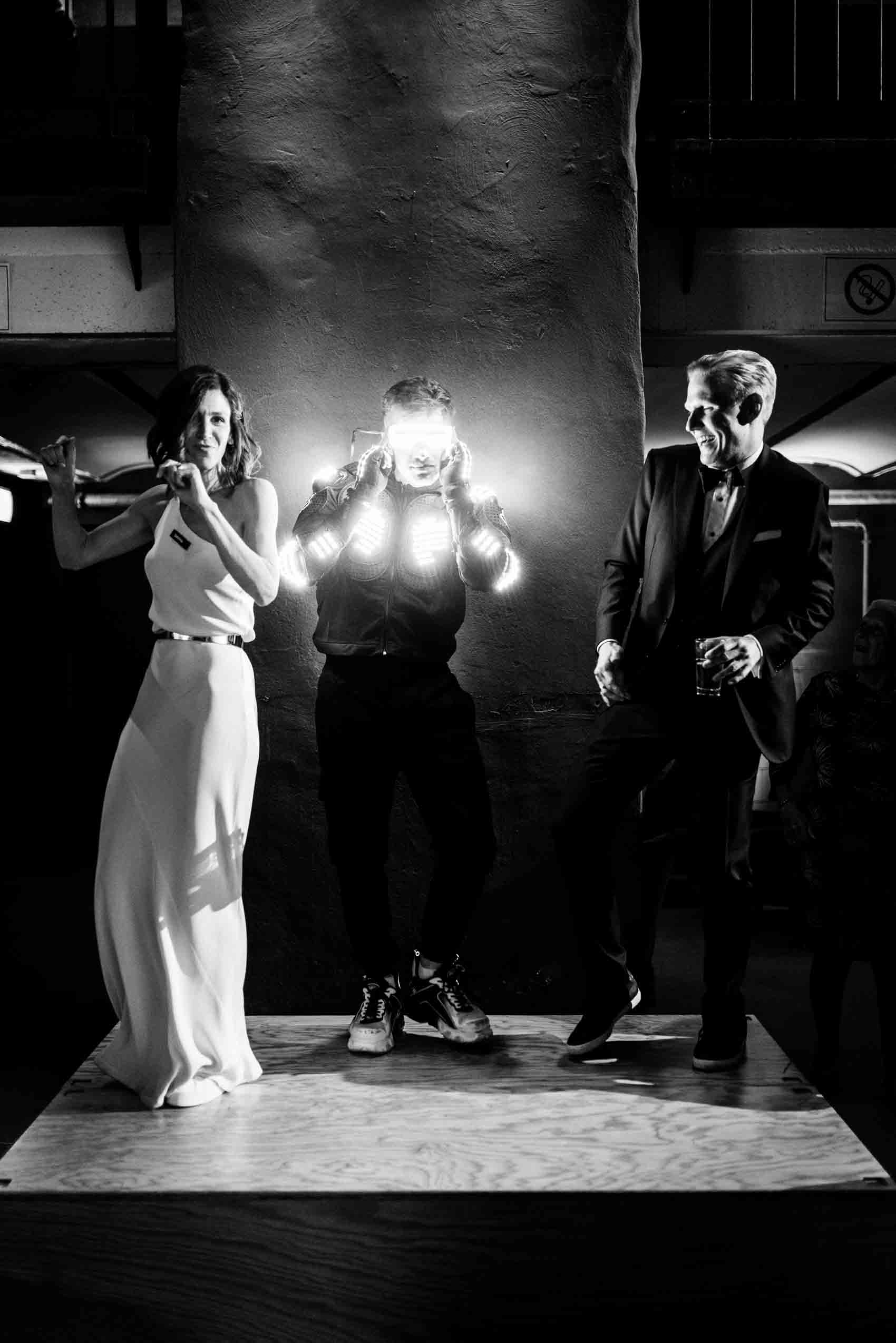 Robodancer tanzt zusammen mit Brautpaar im Club Grey, einem Veranstaltungsraum des Factory Hotel in Münster. Im Vordergrund  eine Ziegelwand sowie jubelnde Hochzeitsgäste zu sehen. 