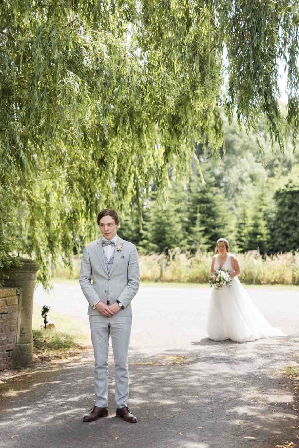 Bräutigam in graugrünem Anzug mit grauer Weste mit Karos wartet beim First Look auf die Braut vor Brücke zu Schloss Buldern in Dülmen