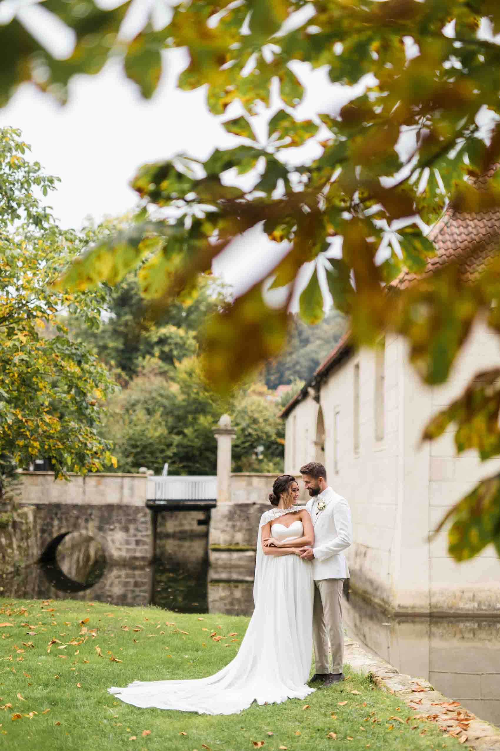 Hochzeitsfoto von Braut mit Cape und Brautkleid mit Schleppe und Bräutigam im weißen Anzug. Das Hochzeitspaar steht vor dem Wasserschloss Haus Marck in Tecklenburg