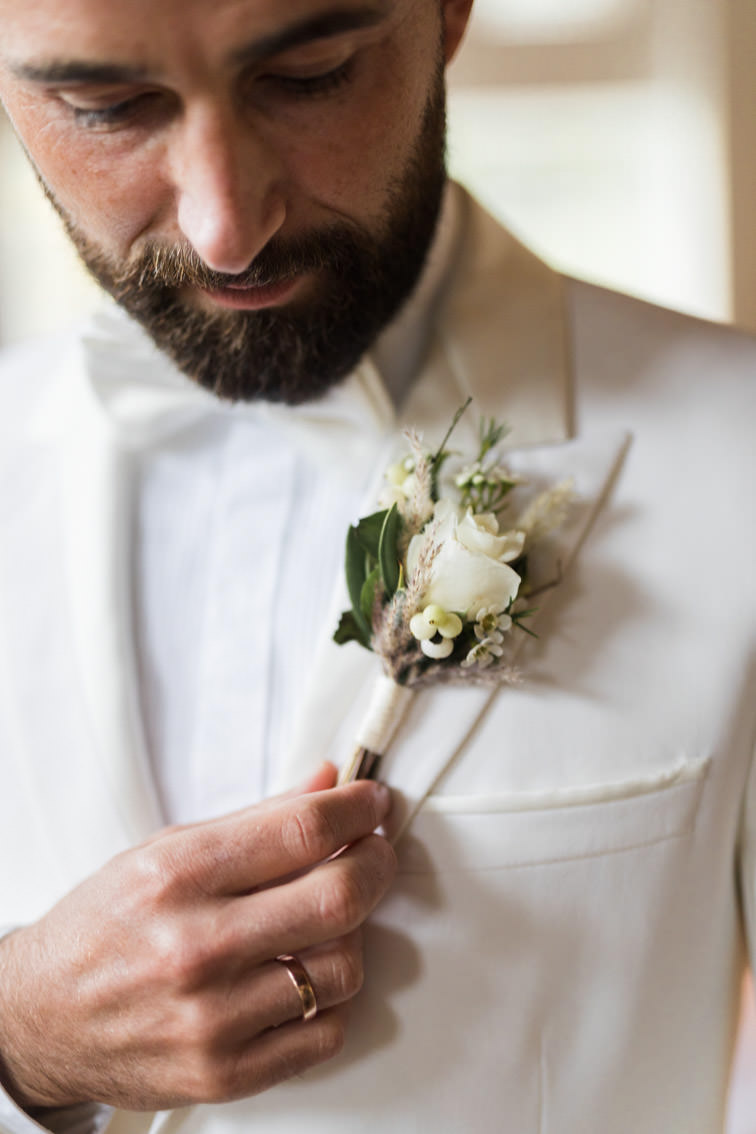 Detailaufnahme eines Hochzeitsansteckers in creme des Bräutigams mit Bart und weißem Anzug