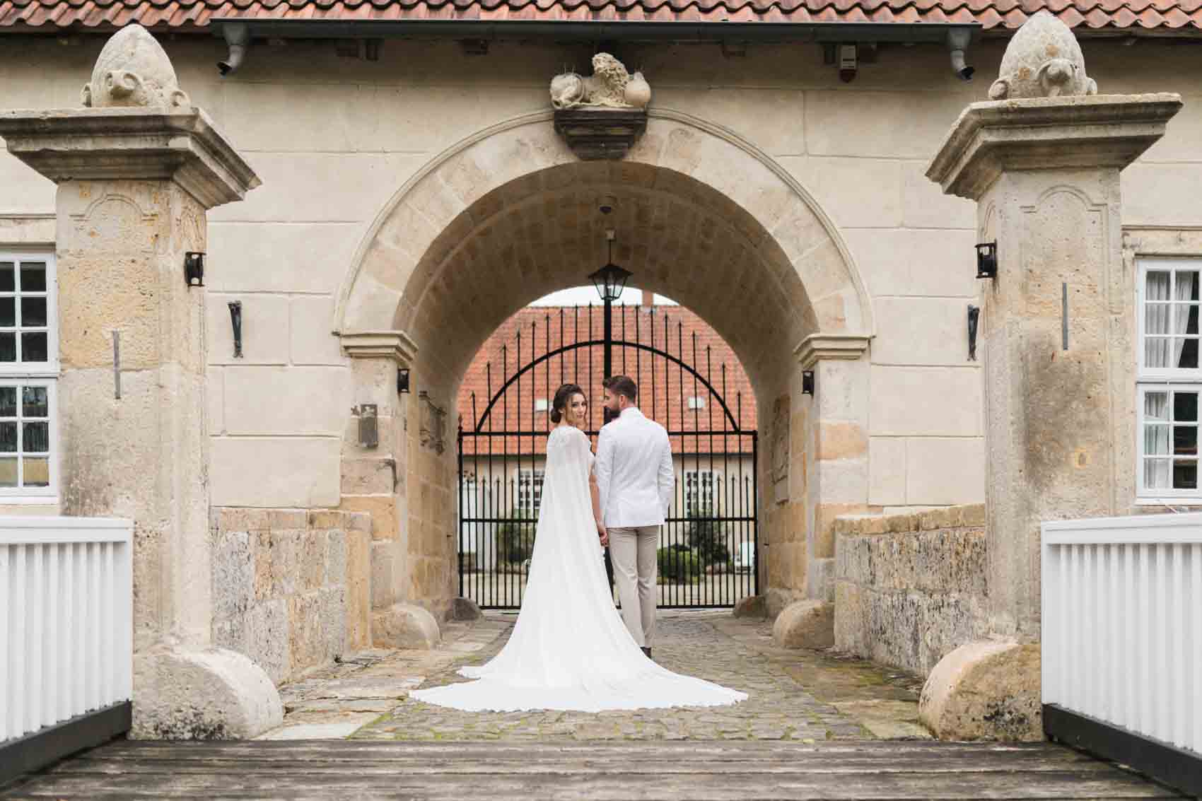 Hochzeitsfoto von Braut mit Cape und Brautkleid mit Schleppe und Bräutigam im weißen Anzug. Das Hochzeitspaar steht vor dem Wasserschloss Haus Marck in Tecklenburg auf der Brücke mit dem Rücken zum Betrachter