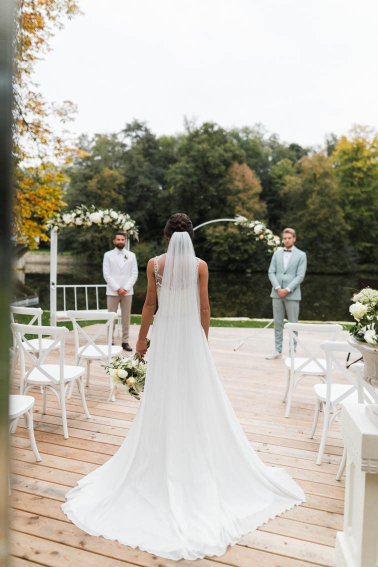 Braut schreitet bei einer freien Trauung auf der Terrasse der Remise von Haus Marck in Tecklenburg auf den Bräutigam zu. Dieser steht vor einem Traubogen mit weißem Blumenschmuck. Der Trauredner Julian Hügelmeyer steht im Hintergrund.