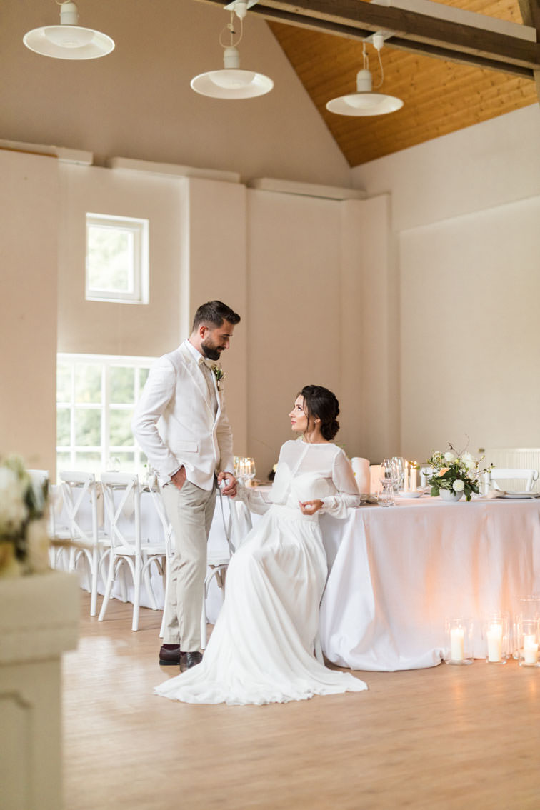 Bräutigam in weißem Anzug steht vor sitzender Braut an einem Gedeckten Hochzeitstisch mit weißen Crossback Chairs in der Remise des Wasserschloss Haus Marck in Tecklenburg.