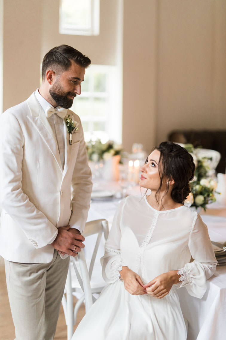 Bräutigam in weißem Anzug steht vor sitzender Braut an einem Gedeckten Hochzeitstisch mit weißen Crossback Chairs in der Remise des Wasserschloss Haus Marck in Tecklenburg.