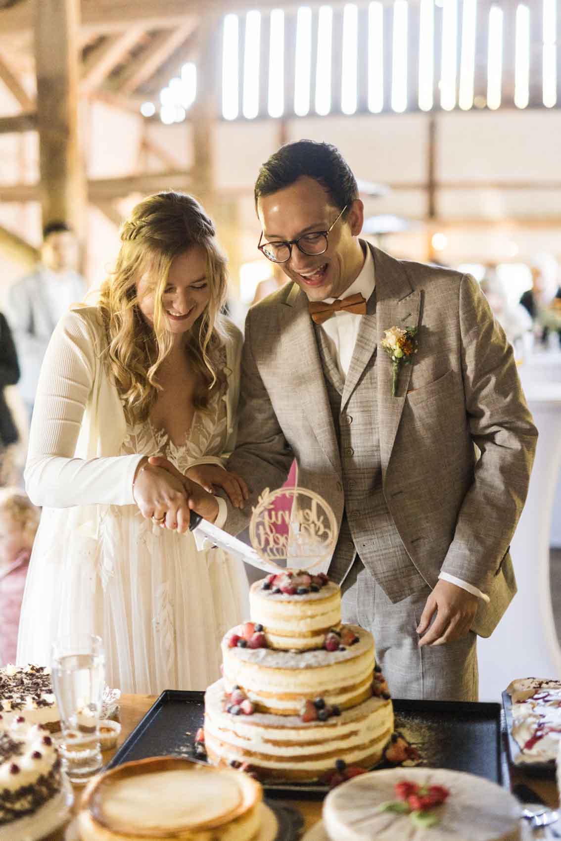 Anschnitt der Hochzeitstorte, einem Naked Cake durch ein Hochzeitspaar in der Scheune der Hochzeitslocation Haus Runde in Billerbeck.