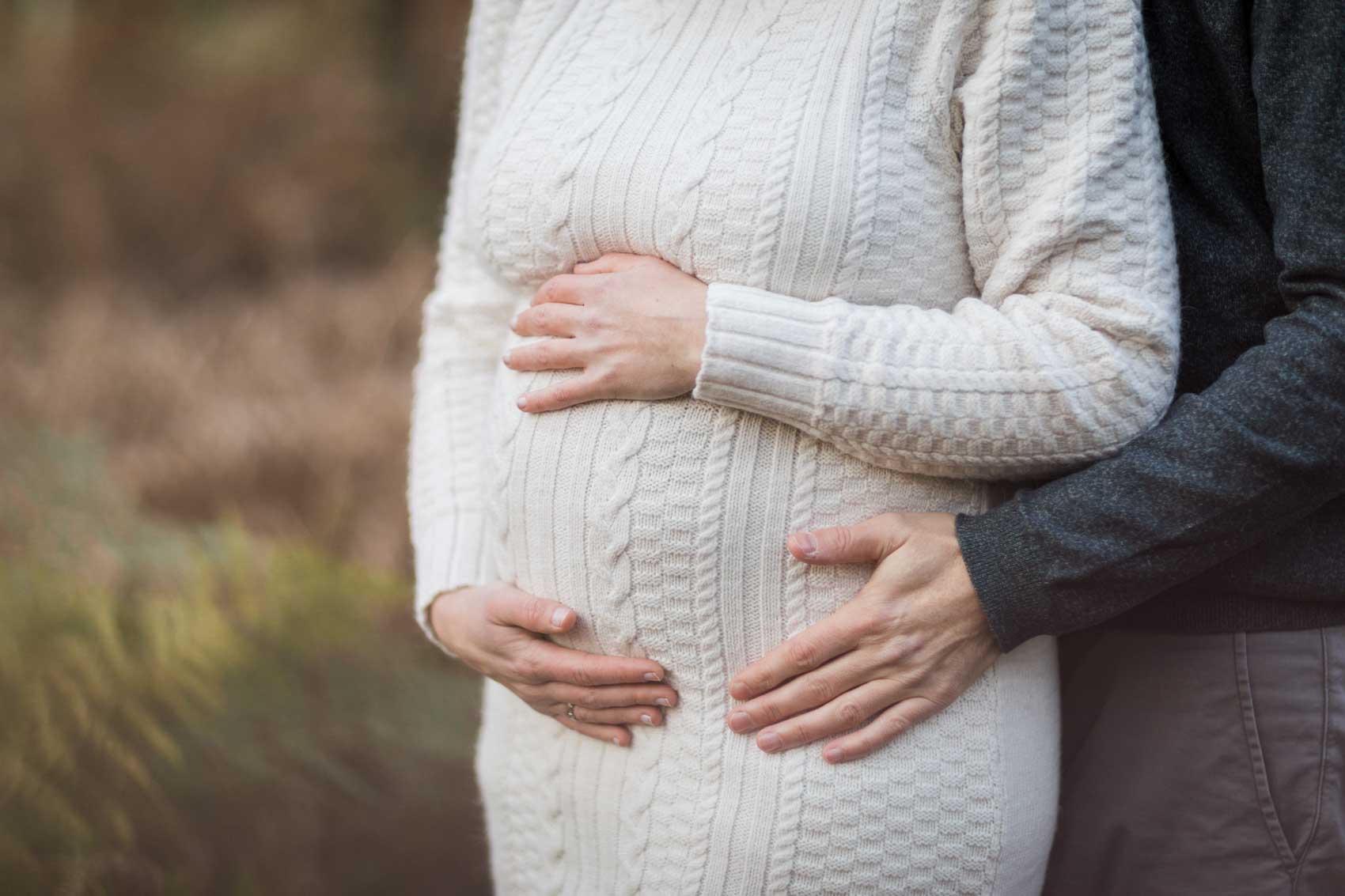 Detailfoto vom Bauch einer schwangeren Frau in einem wollweißen Strickkleid mit Zopfmuster. Die Frau hat beide Hände auf den Bauch gelegt, der Mann hat ebenfalls eine Hand auf dem Bauch seiner Frau