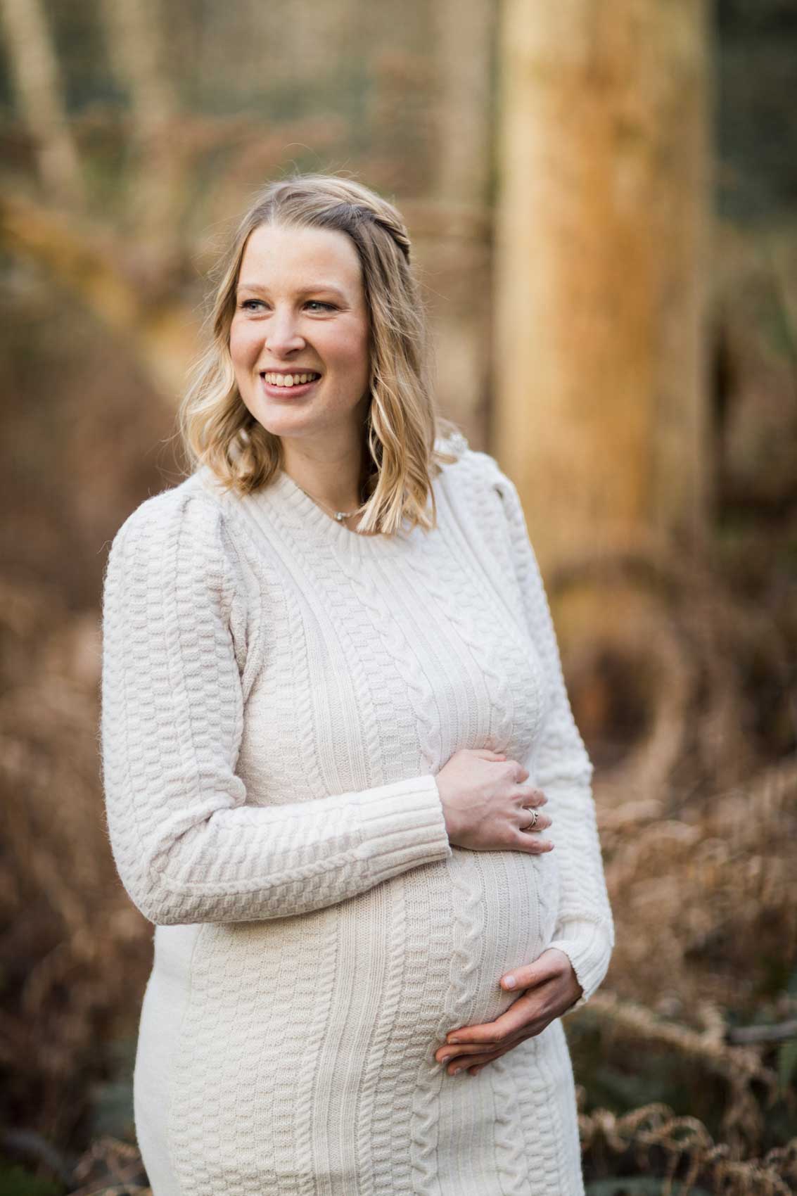 Portraitfoto einer schwangeren Frau in einem wollweißen Strickkleid mit Zopfmuster. Die Frau hat beide Hände auf den Bauch gelegt.