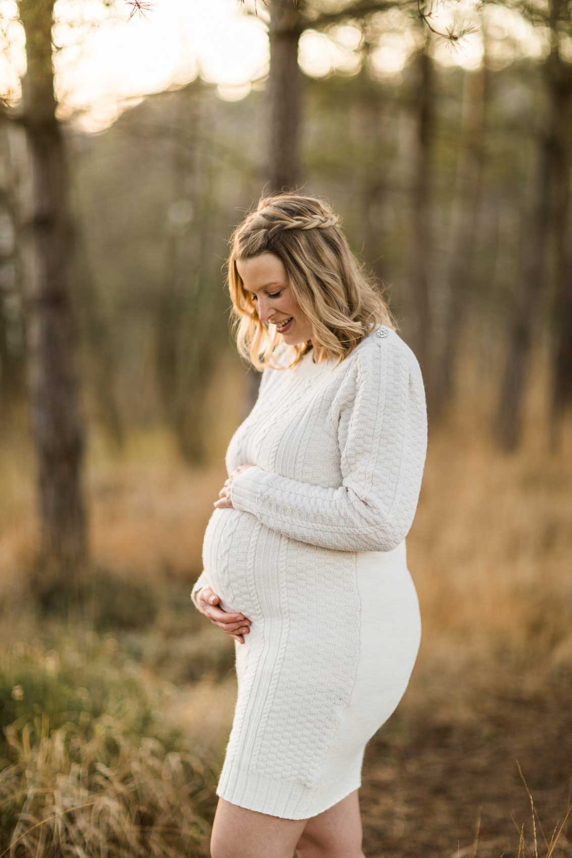 Portraitfoto einer schwangeren Frau in einem wollweißen Strickkleid mit Zopfmuster. Die Frau hat beide Hände auf den Bauch gelegt und schaut hinunter.